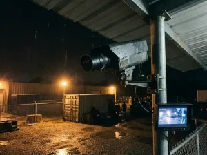 Professionele beveiligingscamera op paal bewaakt industriële binnenplaats bij nacht met regendruppels en amber verlichting