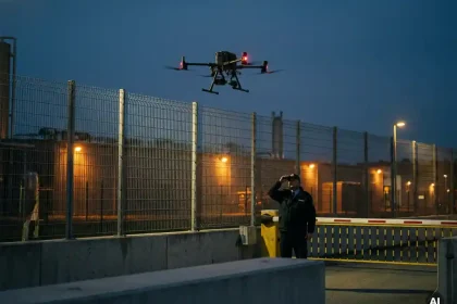 Bewakingsdrone met rode navigatieverlichting boven omheind industrieel terrein bij schemering, bewaker staat bij slagboom.